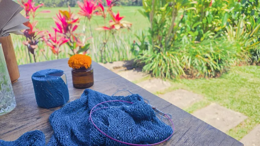 Poncho di jarum bundar dengan benang biru, di atas meja dengan bunga oranye di sampingnya.
