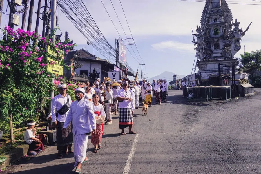 Iringan orang berpakaian upacara putih berjalan di sepanjang jalan, dengan gunung di belakang.